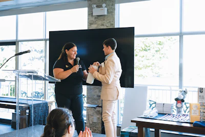 Two women speaking on stage at a professional event with large windows and presentation screen in background