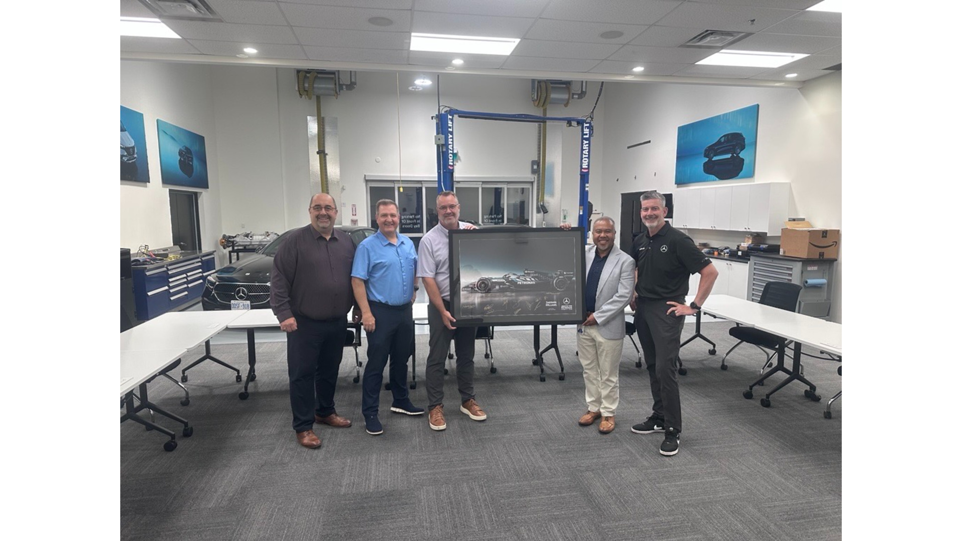 Six men standing together in an industrial workshop displaying a framed photograph, with blue automotive posters on the walls.