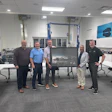 Six men standing together in an industrial workshop displaying a framed photograph, with blue automotive posters on the walls.