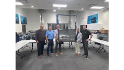 Six men standing together in an industrial workshop displaying a framed photograph, with blue automotive posters on the walls.