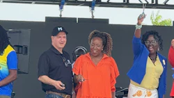 Three people smiling on stage at an outdoor event, wearing colorful clothing including orange, blue, and yellow.