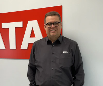 Professional man in glasses and black SATA-branded shirt smiling in front of red SATA company signage