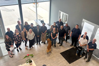 Group of diverse professionals gathered indoors for a team photo, standing together in a modern space with large windows.