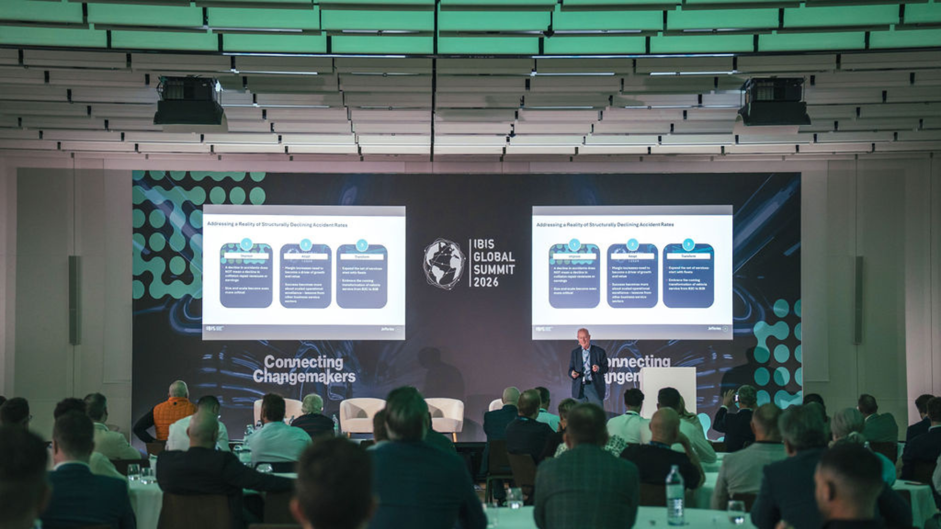 Iris Global Forum conference presentation with speaker addressing audience in modern venue with teal-lit ceiling and dual projection screens