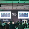 Iris Global Forum conference presentation with speaker addressing audience in modern venue with teal-lit ceiling and dual projection screens