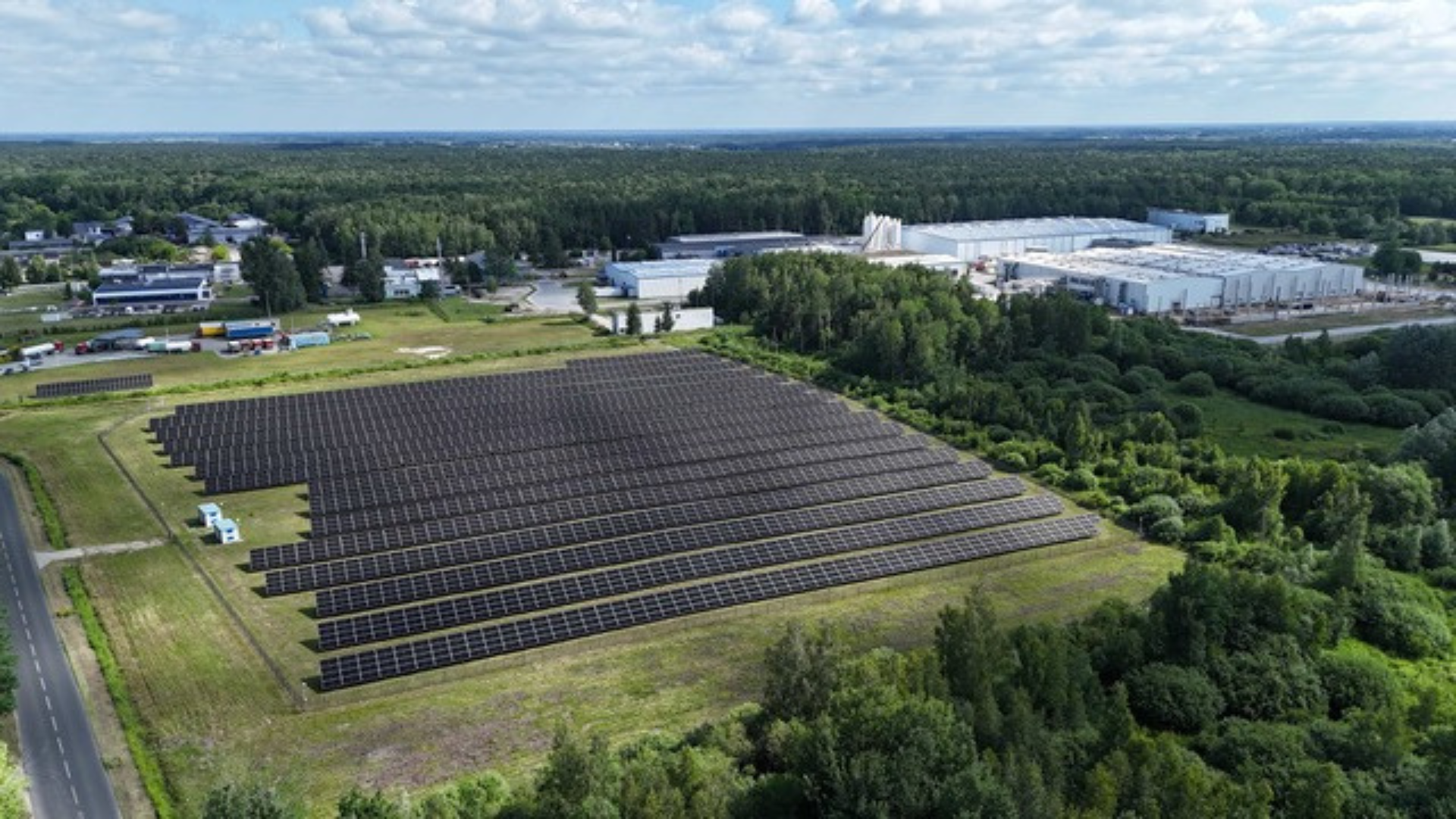 Aerial view of large solar panel farm adjacent to industrial manufacturing facilities surrounded by forest