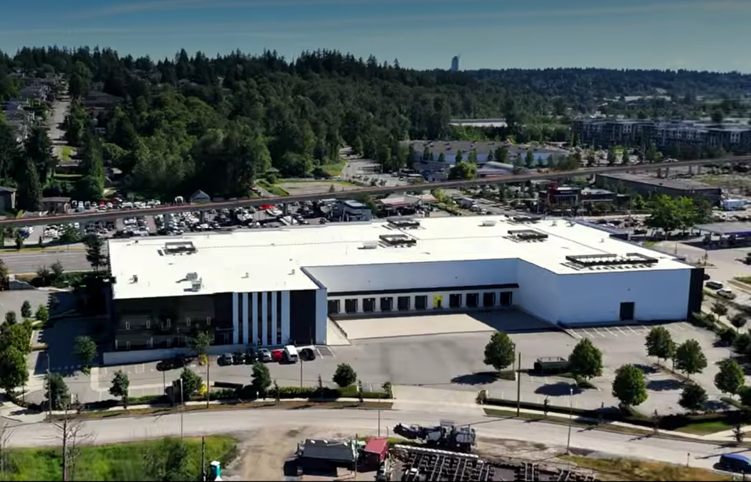 Aerial view of modern industrial building with white roof and dark facade surrounded by parking lots and forested landscape