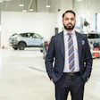 Professional man in dark suit and striped tie standing in modern automotive service facility with vehicles in background