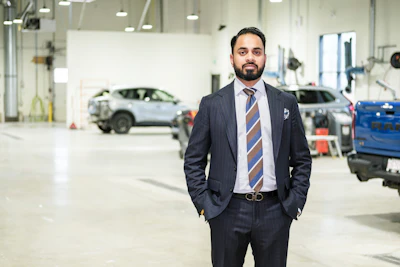 Professional man in dark suit and striped tie standing in modern automotive service facility with vehicles in background