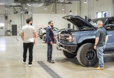 Three men examining a blue Ford Bronco with open hood in a modern vehicle showroom with other vehicles visible in background.