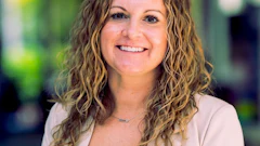 Professional headshot of a woman with wavy brunette hair smiling at camera wearing a light beige blazer