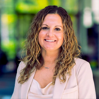 Professional headshot of a smiling woman with long wavy brown hair wearing a beige blazer