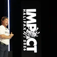 Speaker presenting at IMPACT Halfax 2026 conference with large event branding signage and blue accent lighting