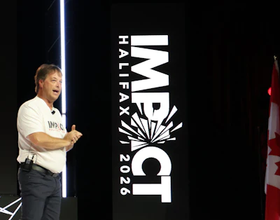 Speaker presenting at IMPACT Halfax 2026 conference with large event branding signage and blue accent lighting
