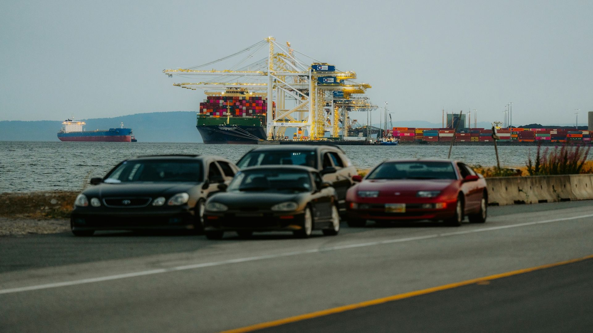 Three parked cars at a busy cargo port with container ships and yellow loading cranes in the background