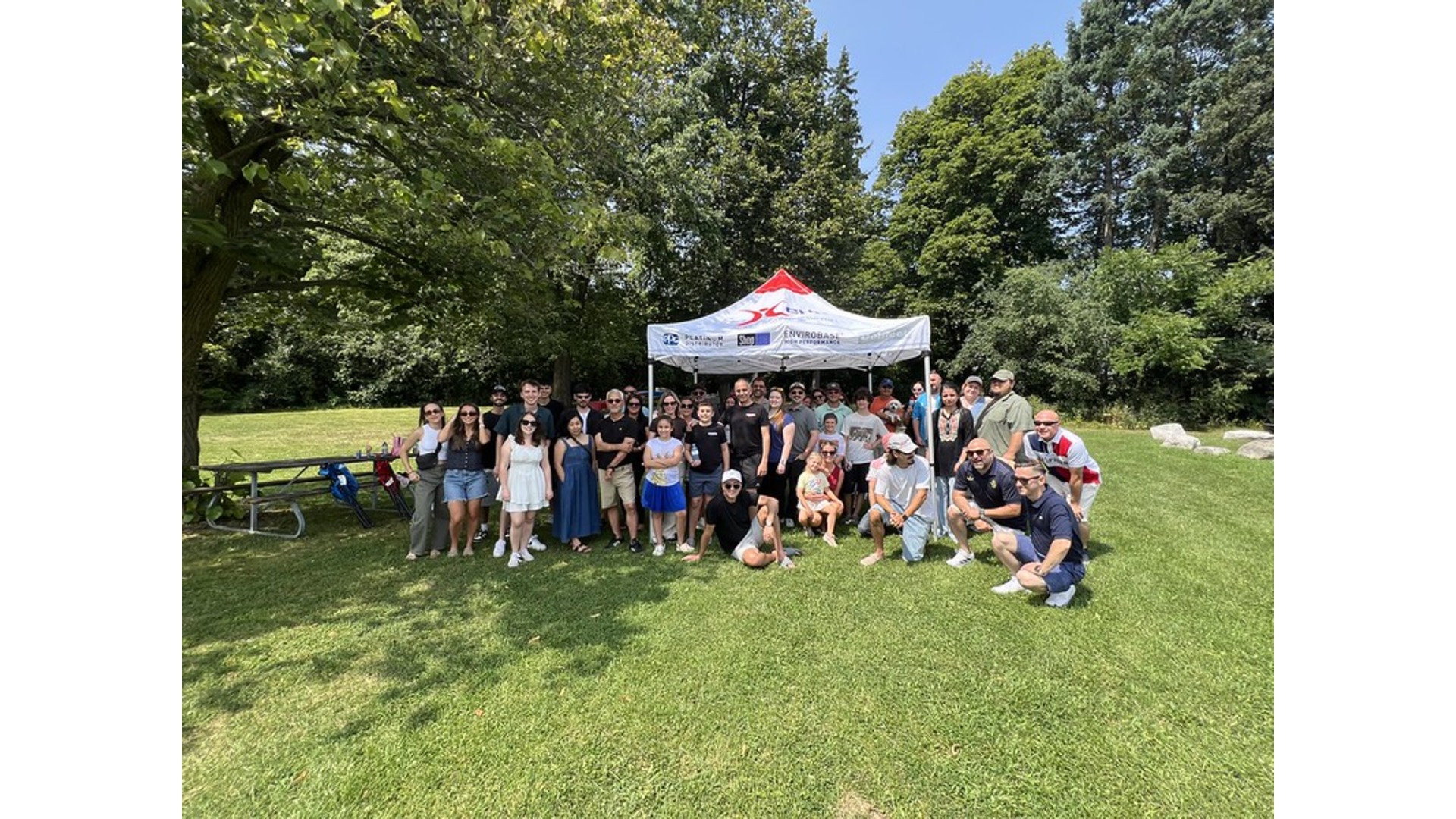 Large group of people posing together under a white tent at an outdoor park event on a sunny day