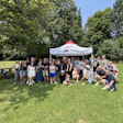 Large group of people posing together under a white tent at an outdoor park event on a sunny day