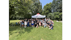 Large group of people posing together under a white tent at an outdoor park event on a sunny day