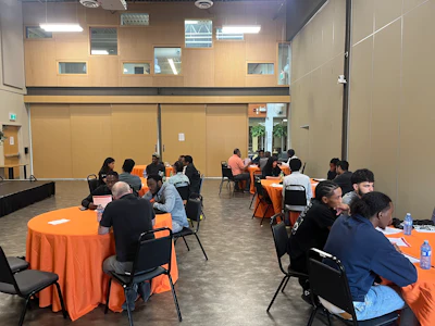 Group of people seated at orange-clothed round tables in a modern event venue with beige walls and skylights.