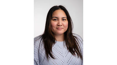 Professional headshot of a woman with long dark hair wearing a light purple textured sweater against a neutral gray background