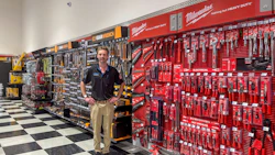Man standing in tool store showroom with red Milwaukee tool display wall and black and yellow DeWalt pegboard tool organization system.