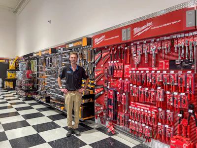 Man standing in tool store showroom with red Milwaukee tool display wall and black and yellow DeWalt pegboard tool organization system.