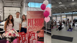 Three staff members stand at a Pit Stop Pilates promotional event in an automotive showroom with pink balloons and flowers.