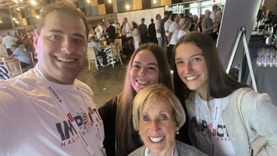 Four smiling people wearing branded event t-shirts at an indoor gathering with attendees visible in the background
