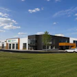 Modern commercial building with black and white facade and yellow accents, set on green lawn with parked vehicles and young trees