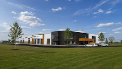 Modern commercial building with black and white facade and yellow accents, set on green lawn with parked vehicles and young trees