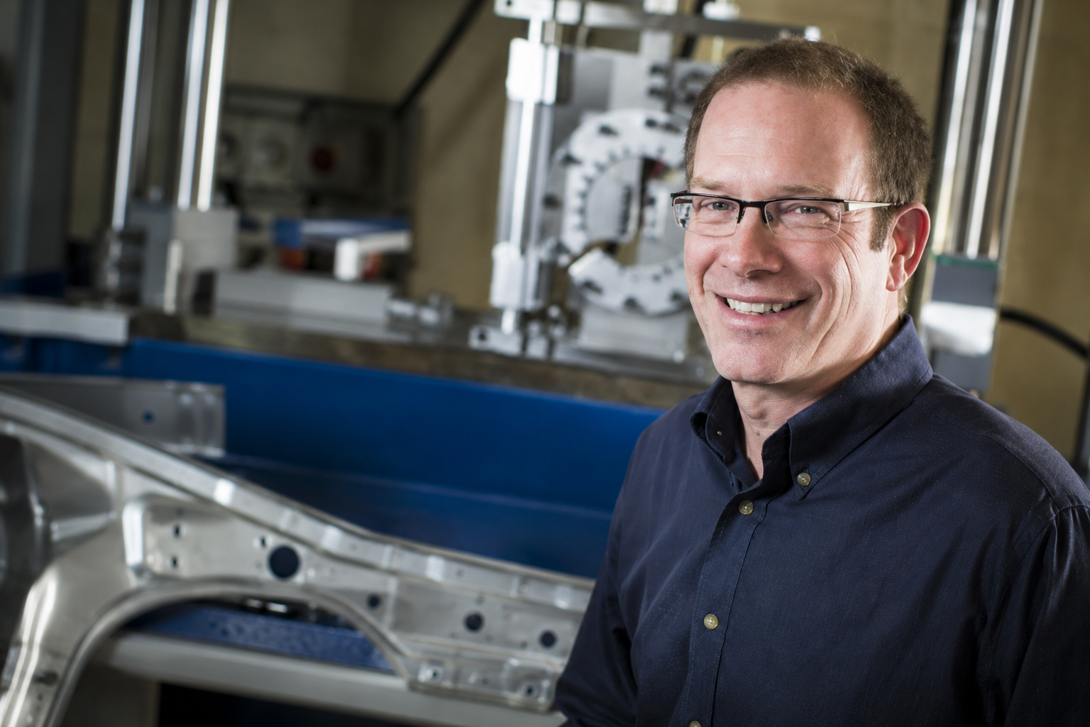 Professional man wearing glasses and navy shirt smiling in front of industrial manufacturing equipment