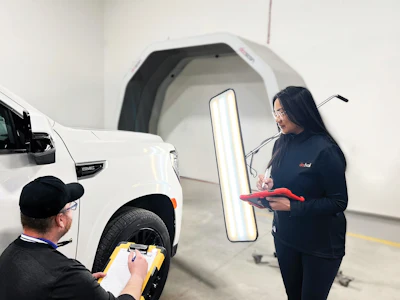 Technician inspecting white vehicle door with measurement tool while colleague takes notes during quality control inspection