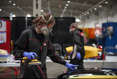 Worker wearing protective gear and blue gloves sanding a vehicle surface in an industrial workshop with equipment and other workers visible in background