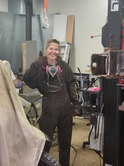 Smiling woman wearing black hoodie and pink heart-shaped headphones in a workshop studio surrounded by materials and equipment