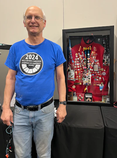 Man wearing blue Skills Ontario 2024 Competition t-shirt standing next to display case with red jacket and pins
