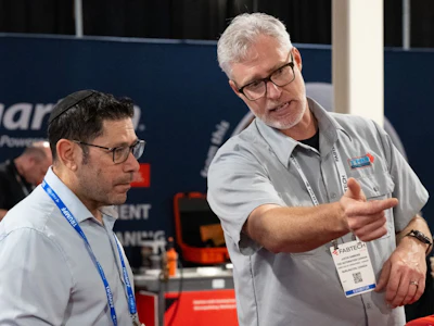 Two professionals wearing conference badges discuss at an indoor event, with exhibition signage visible in the background.
