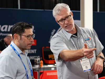 Two professionals wearing conference badges discuss at an indoor event, with exhibition signage visible in the background.