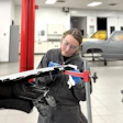 Technician in black jacket inspecting vehicle hood with red tool in automotive repair facility