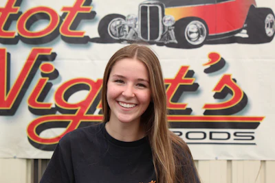 Smiling young woman in black shirt standing in front of Hot Nights Rods vintage car shop signage