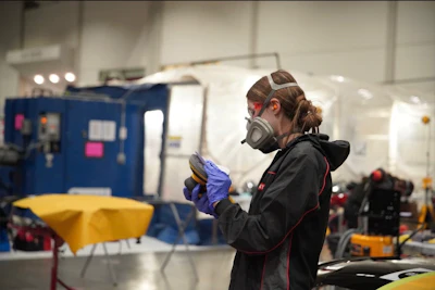Worker wearing safety mask and gloves examining materials in industrial workshop with blue equipment in background