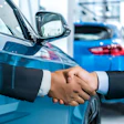 Two businesspeople shake hands in front of blue vehicles at an automotive dealership showroom