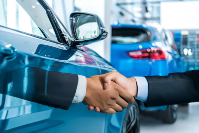 Two businesspeople shake hands in front of blue vehicles at an automotive dealership showroom