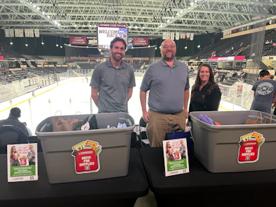 Three people hold collection bins with promotional materials at an ice hockey arena