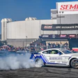 White and blue Shelby drift car performing at SEMA motorsports event with crowd and sponsor banners