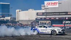 White and blue Shelby drift car performing at SEMA motorsports event with crowd and sponsor banners