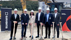 Six professionals in hard hats hold shovels at groundbreaking ceremony with excavator and branded signage