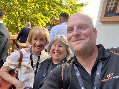Three smiling people at an outdoor event, with one wearing a BodyShop Connect branded polo shirt