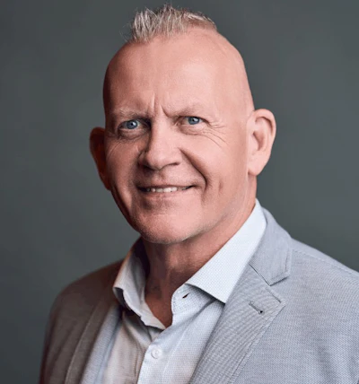 Professional headshot of a smiling middle-aged man in light blue button-up shirt against gray background
