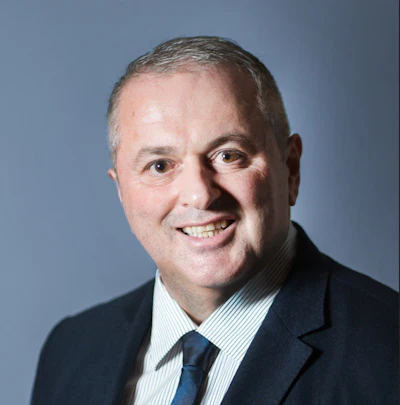 Professional headshot of a smiling man in dark business suit and blue tie against blue background