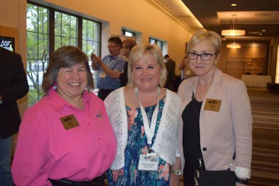 Three women smiling at a professional conference event, wearing name badges and lanyards in an indoor venue.